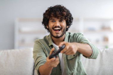Young happy indian man laughing and playing video games on weekend, sitting on couch alone at home, holding joystick, having fun, copy space. Modern technologies and entertainment concept