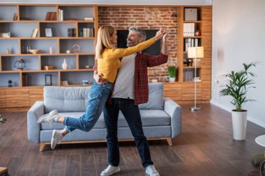 Satisfied adult european man and woman in casual dancing in new house, have fun, enjoy relaxing in living room interior, free space. Spare time in cozy own home, love, romance and family relationship
