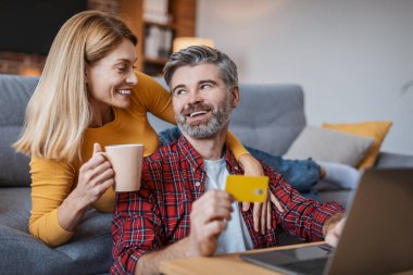 Smiling adult european woman with cup of drink hug man with laptop and credit card, enjoy shopping online remotely, sit on sofa in room interior. Order, money payment, shopaholics and sale at home