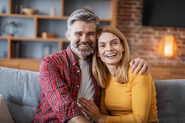Smiling middle aged handsome european male and female hugging and enjoy tender moment, look at camera, sitting on sofa in living room interior. Love, family relationships at home, people emotions