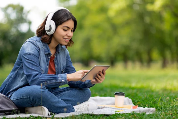 Young Arab Woman In Wireless Headphones Using Digital Tablet While Relaxing Outdoors, Middle Eastern Female Student Sitting On Lawn In Park And Browsing App On Modern Gadget, Copy Space