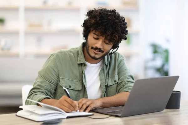 Young indian man having online training or attending webinar, sitting at workdesk, using laptop and headset, taking notes and smiling, home interior, copy space. Online education concept