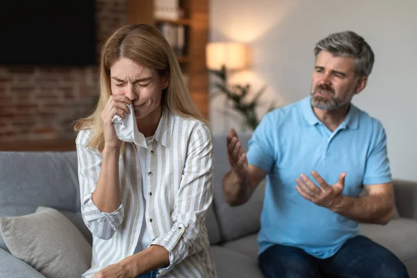 Angry upset mature european husband yells at crying wife, couple quarreling in living room interior. Human emotions, relationship problems at home, stress, pressure, scandal and domestic violence