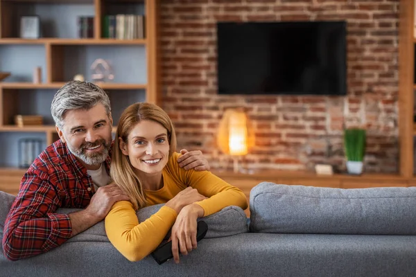 Happy adult european man and woman looking at camera, enjoy relaxing in living room interior with tv with blank screen. Free time in cozy home, love and relationships, news, movies in evening together