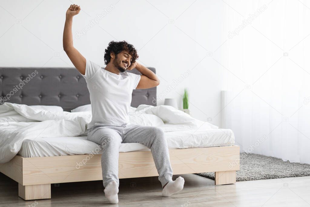Cool attractive millennial indian guy stretching in bed at home, young man waking up in white bedroom, wearing pajamas, spending weekend alone, copy space. Leisure, resting, domestic lifestyle