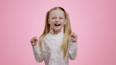 Cute happy little girl with two ponytails enjoying success or present, laughing and gesturing, pink studio background, slow motion, free space