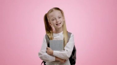 Back to school. Adorable little girl pupil with backpack carrying books, smiling to camera, pink studio background, slow motion