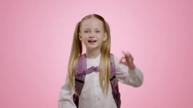 Adorable little schoolgirl with backpack showing Ok gesture, approving great kids stuff and content, pink studio background, slow motion