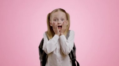 Amazing news. Cute surprised little schoolgirl with backpack enjoying unbelievable information, touching her cheeks in amazement, pink studio background, slow motion