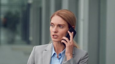 Worldwide mobile connection. Close up portrait of serious middle aged business lady talking on smartphone with partners or family, standing near office building, slow motion