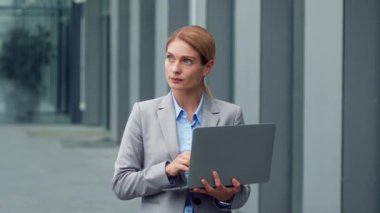 Business on go. Confident professional businesswoman typing on laptop, standing outdoors near office building, slow motion, free space