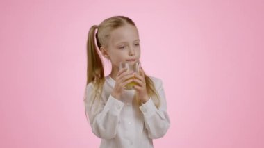 Healthy drink and vitamins. Cute little girl drinking fresh orange juice from glass, enjoying citrus taste, smiling over pink studio background, slow motion, free space