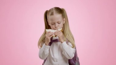 Tasty lunch. Cute little schoolgirl with backpack eating homemade sandwich, enjoying homemade snack during break, pink studio background, slow motion