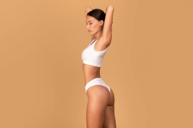 Beautiful slim woman posing in white underwear with hands behind her head over beige studio background, showing perfect slender body, copy space