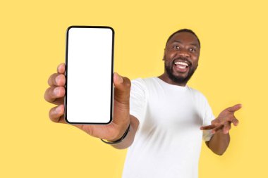 Cheerful Black Man Showing Huge Smartphone With Empty Screen To Camera On Yellow Studio Background. Check This Mobile Application Concept. Selective Focus, Mockup