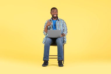 Happy Black Male Using Laptop Computer Gesturing Thumbs Up Approving Online Service Sitting Over Yellow Studio Background. Guy Showing Like Gesture Browsing Internet. Full Length