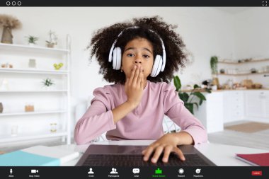 Surprised african american kid cute school girl with nice bushy hair sitting at desk in front of laptop at home, using wireless headset, having video call with friend, touching her mouth, screenshot