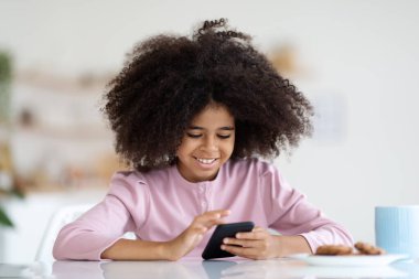 Cool african american child using cell phone at home, happy black school girl sitting at table, drinking juice, eating cookies and chatting with friends on smartphone via mobile app, copy space