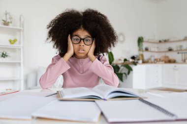 Overwhelmed african american school girl with bushy hair sitting at desk full of books, notepads, stressed kid pupil doing homework alone, wearing eyeglasses, copy space. Difficulties with education