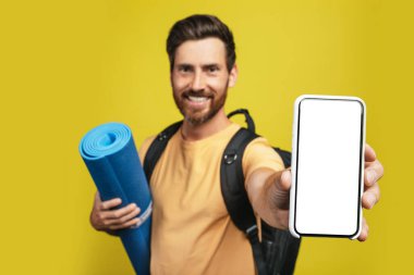 Online travel agency. Excited male tourist showing smartphone with blank screen and holding touristic mat over yellow studio background. Mockup for website or ap design