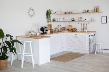 Light stylish kitchen in daylight, simply, minimalist scandinavian interior. White furniture, modern coffee machine, small wooden shelves, green plants in pots