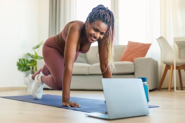 Young african american fitness lady doing leg exercises and watching online workout on laptop at home. Sports lifestyle and online lesson with trainer