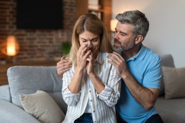 Sad middle aged caucasian man with beard hug, calm crying unhappy wife in living room interior. Family support at home, relationship problems, people emotions, depression and stress due covid-19