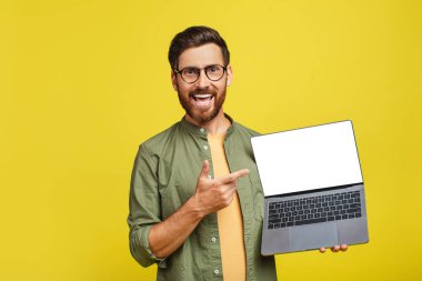 Happy middle aged man pointing at empty laptop screen with space for your design over yellow studio background, mockup. Stylish guy showing PC with space for your advertisement