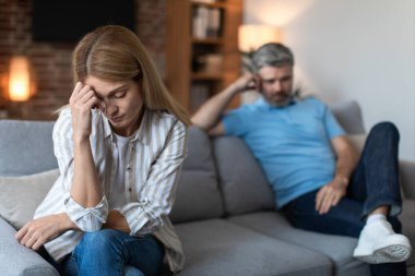 Upset middle aged caucasian male with beard ignores offended lady after conflict in living room interior. Quarrel, relationships problems, people emotions, depression, disagreement and stress at home
