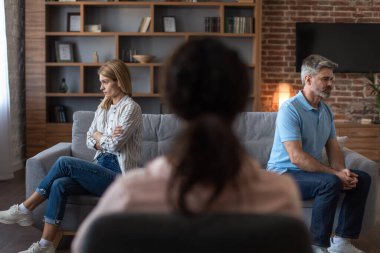 Offended angry adult caucasian man ignores woman at meeting with black psychologist in office clinic interior, back. Doctor help and family therapy, relationship problems, quarrel and consultation