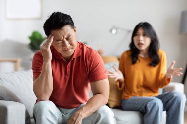 Emotional asian spouses having fight at home, angry chinese husband and wife sitting on couch, yelling and gesturing, having difficulties in marriage. Divorce, breakup, crisis in relationship