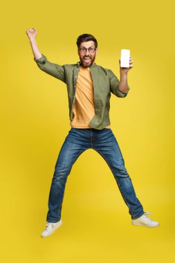 Yes, cool mobile offer. Joyful man showing empty cellphone screen and jumping up in the air on yellow studio background. Happy male recommend website, copy space