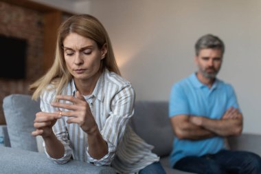 Offended sad adult caucasian woman takes off ring and ignores unhappy husband at conflict in living room interior. Problems in relationships, emotions after quarrel at home, divorce and breakup