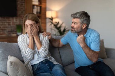 Angry aggressive mature caucasian husband with beard threatens to scared wife with fists in living room interior. Problems in relationships, emotions, quarrel at home, beating and domestic violence