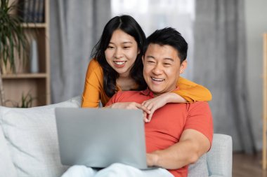 Positive asian couple sitting on sofa, using computer at home, happy chinese lady hugging her boyfriend, looking at laptop screen and smiling, websurfing together, copy space