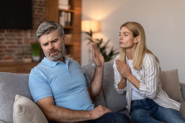 Adult caucasian wife asks and prays with gesture at sad husband with beard, man ignores lady in living room interior. Problems in relationships, people emotions at home, desires, hope and apologize