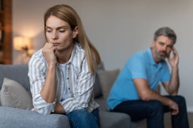 Unhappy depressed mature caucasian husband with beard ignores offended wife in living room interior. Conflict, family crisis, psychological abuse, relationship problems at home and stress due covid-19