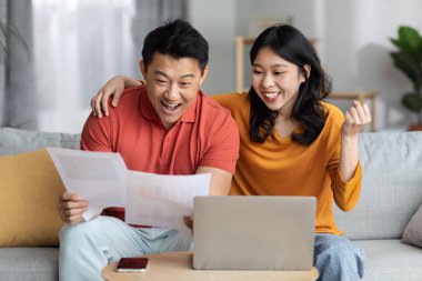 Emotional asian spouses sitting on couch, using laptop, reading correspondence, happy chinese man and woman holding papers and celebrating success, got approved for mortgage, copy space