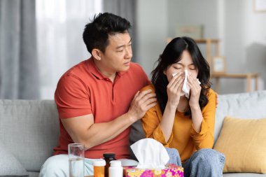 Loving asian husband taking care of sick wife sitting on sofa next to tea table full of medicine and sneezing nose, loving man hugging his ill girlfriend. Cold, flu, coronavirus concept