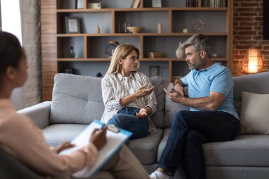 Offended sad nervous adult caucasian husband and wife swear at meeting with psychologist in office clinic interior. Relationship problems, quarrel and human emotions, doctor help and family therapy