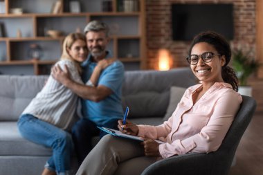 Happy adult caucasian husband hug wife at meeting with psychologist in office clinic interior with young black doctor. Relationship problem solving, reconciliation, medical help and family therapy