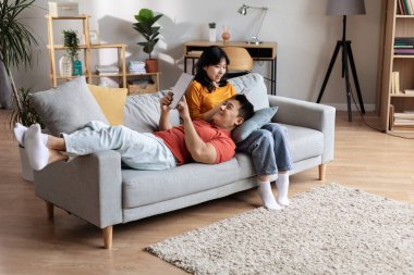 High angle view of beautiful chinese couple relaxing at home, joyful asian man lying on his girlfriend or wife lap, using digital tablet, young woman checking social media on cell phone, copy space