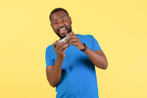 Excited Black Guy Playing Mobile Game On Cellphone Having Fun Standing Over Yellow Studio Background. Male Gamer Using Phone. Gaming Hobby And Leisure Concept