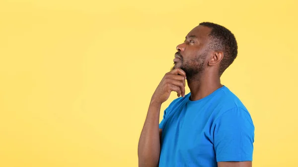 Side View Of Pensive Black Man Thinking Touching Chin Looking Aside At Copy Space Standing Over Yellow Studio Background. Guy Advertising Offer. Let Me Think. Panorama