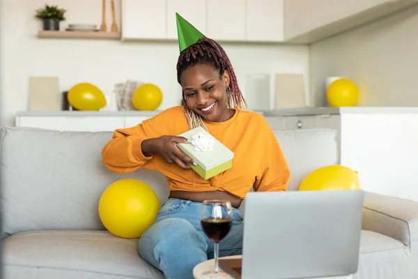 Happy african american lady looking at laptop receiving gift from friends, celebrating b-day via video call at home. Virtual holiday celebration and presents delivery concept