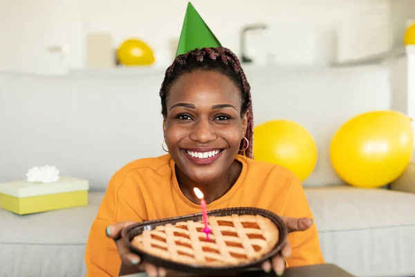 Happy birthday to you. Happy african american lady in festive hat holding pie with burning candle and smiling to camera, sitting at home. Holiday celebration concept
