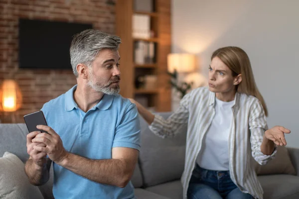 Sad angry middle aged caucasian woman yelling at husband, gesticulates, man suffers from gadget addiction and does not give phone back in living room interior. Quarrel at home, relationship problems