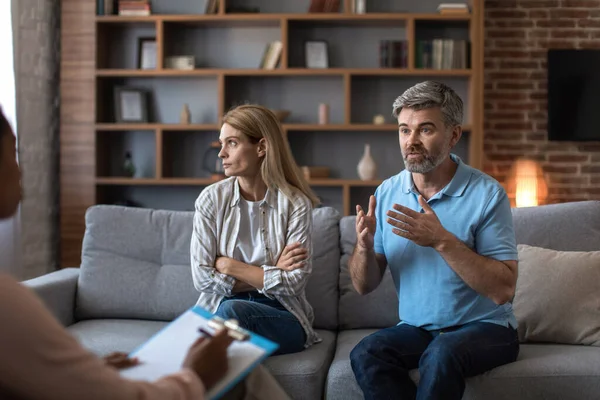 Offended mature caucasian woman ignores man with beard at meeting with psychologist in office clinic interior. Quarrel, doctor help and family therapy, relationship problems and medical support