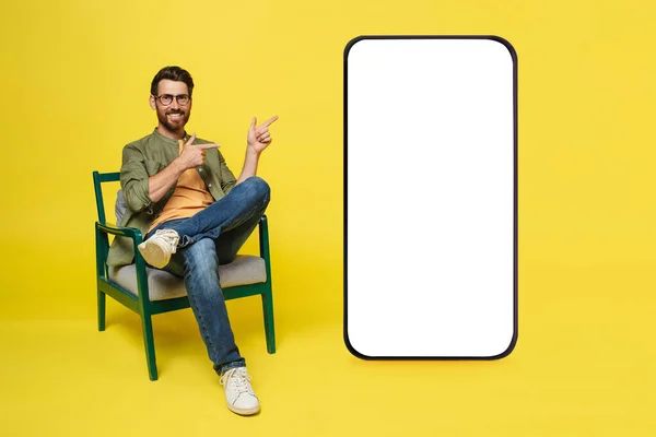 Happy man sitting in armchair and pointing at giant cellphone with empty white screen, mockup for online ad, mobile app or new website design. Smartphone template