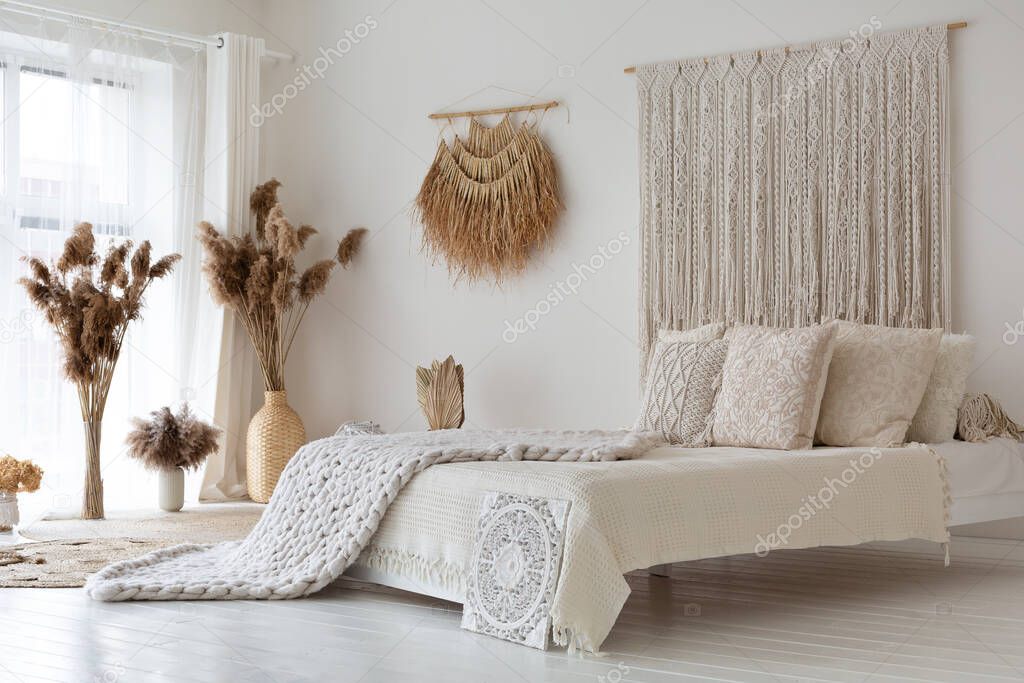 Empty white bedroom full of day light made in original ethnic style, room decorated with straw elements, dry plants, knitted details, big comfortable bed full of beautiful pillows